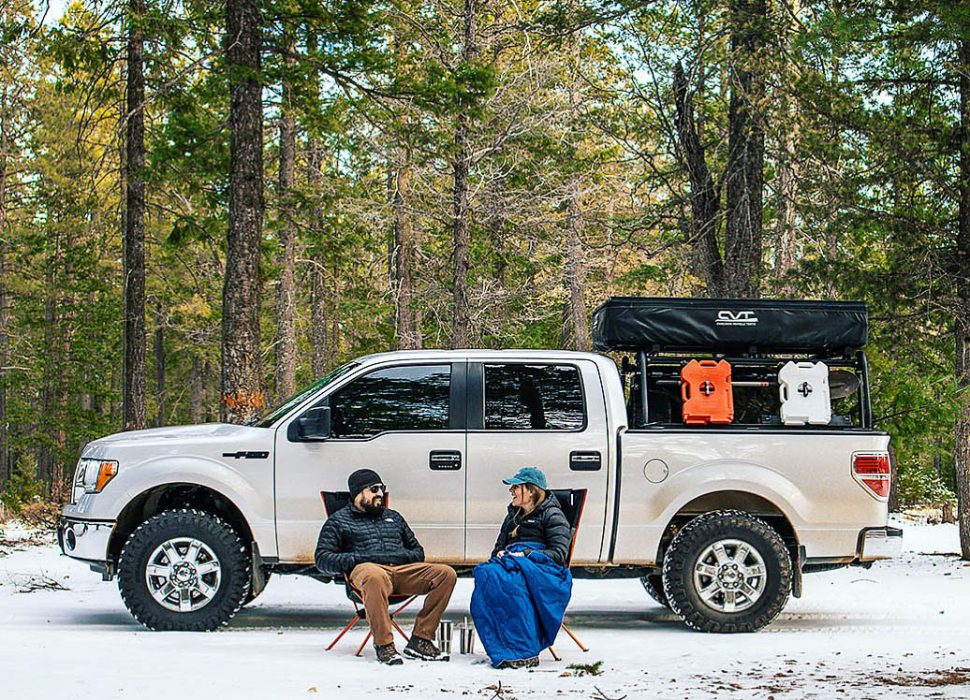 Couple sitting in front of 4x4 truck in chairs on the snow with pine trees behind